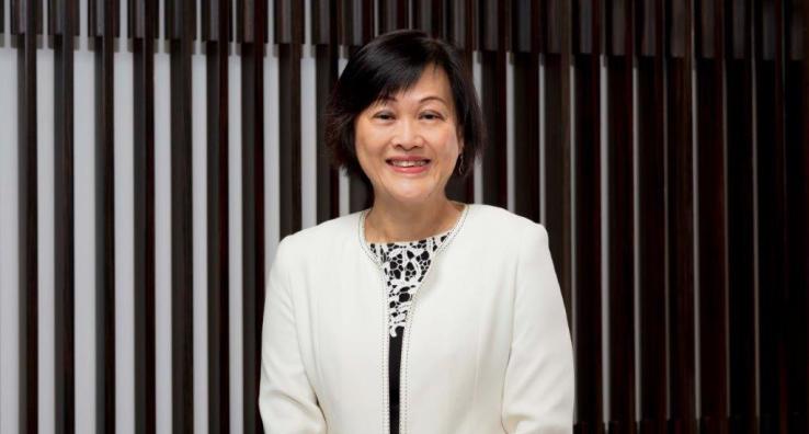 May Tan stands indoors wearing a white jacket, smiling