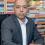 A man wearing a blazer looking at the camera in front of a shelf of books