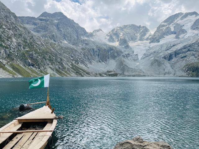 A small wooden boat on the shore of a lake surrounded by snowy mountains in Pakistan. The Pakistani flag flies at the front of the boat.