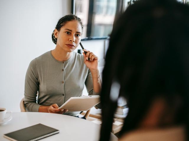 A woman wearing a gray sweater sits, listening to someone in the foreground