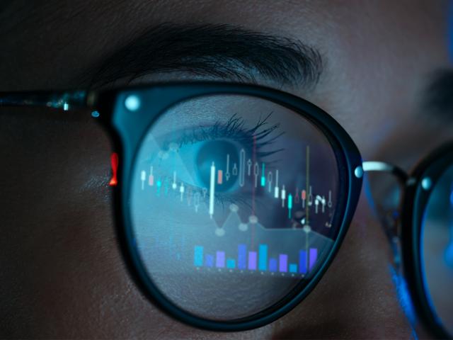 Close up of a person wearing glasses. A blue computer screen with digital graphs in reflected in their glasses.
