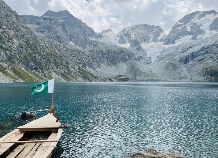 A small wooden boat on the shore of a lake surrounded by snowy mountains in Pakistan. The Pakistani flag flies at the front of the boat.