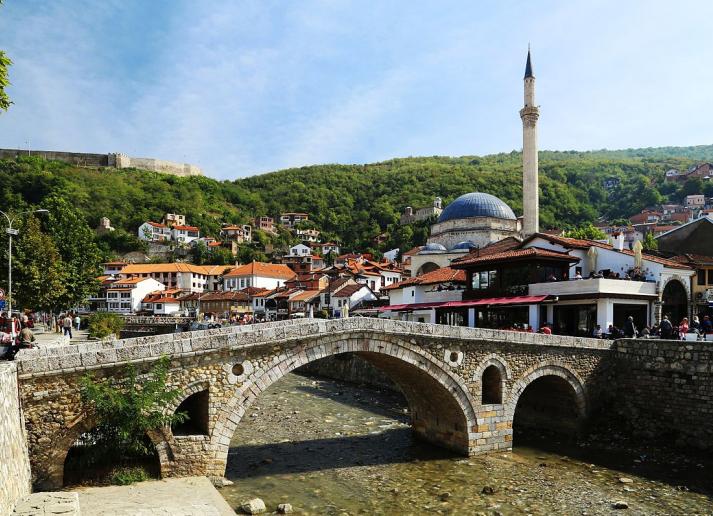 A stone bridge over a creek in a village with a stone tower and red roofs
