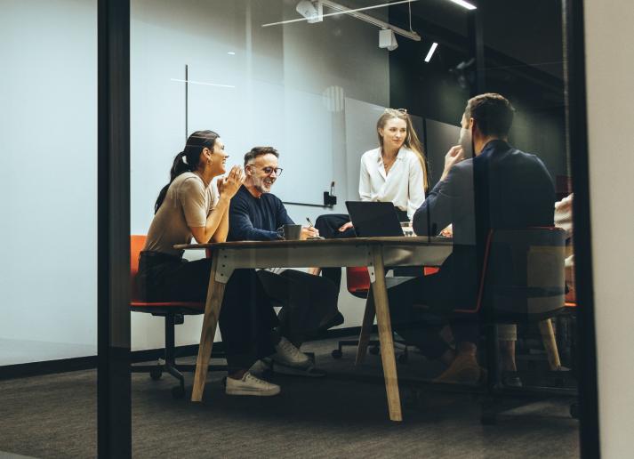 Four office workers in casual attire talk around a table in a collaborative workspace