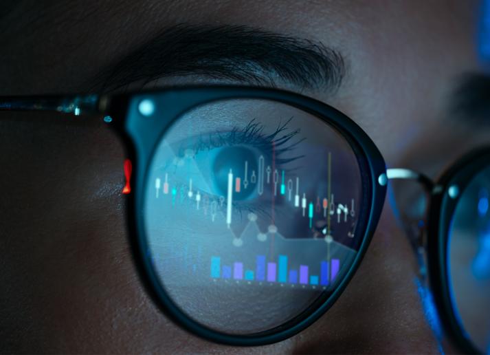 Close up of a person wearing glasses. A blue computer screen with digital graphs in reflected in their glasses.