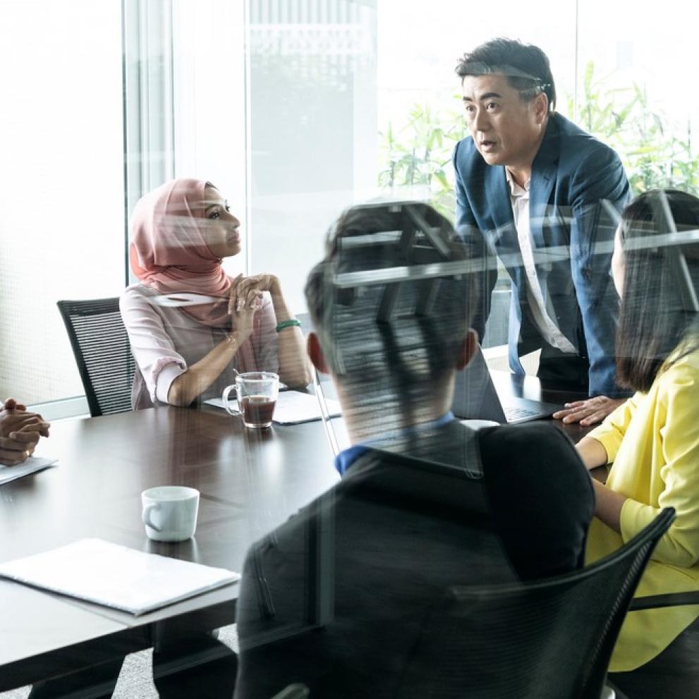 Man standing, leaning on table of colleagues, speaking