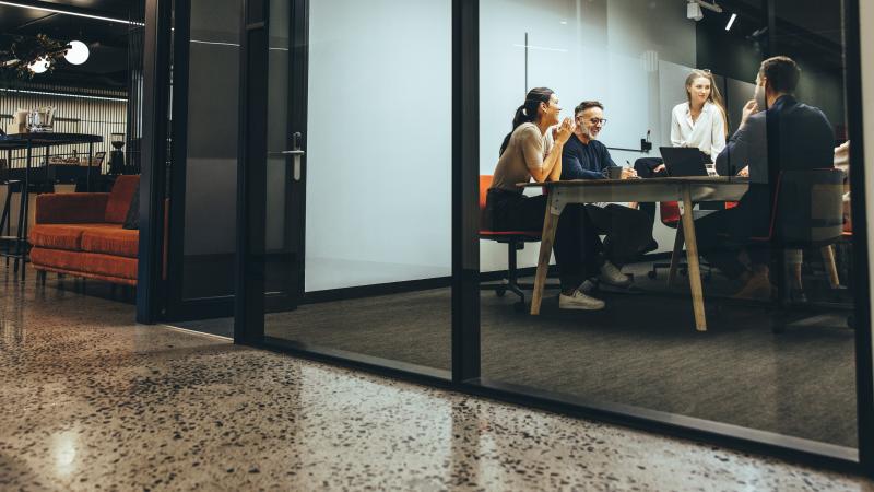 A wide view of a spacious office. Four colleagues in casual attire talk around a table in a collaborative workspace.