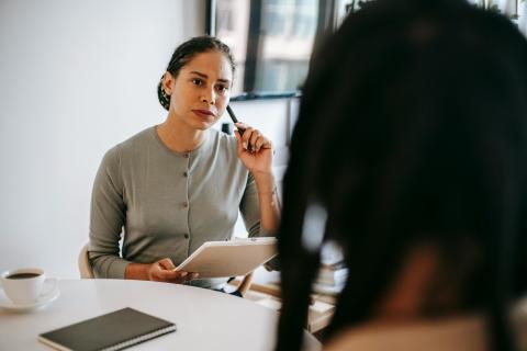 A woman wearing a gray sweater sits, listening to someone in the foreground