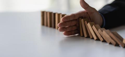 A man's hand stops wooden dominoes from falling into each other on a white table.