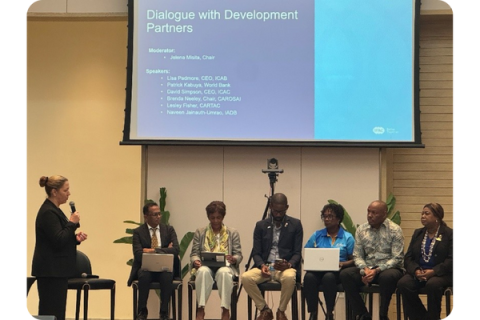 A woman stands with a microphone, speaking to a number of people on stools