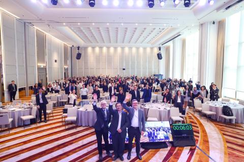 Three event speakers wave at the camera in front of the audience of an event